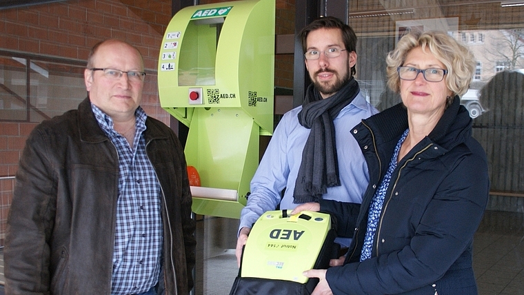 Othmar Zimmermann (links) hat den Kasten per Knopfdruck geöffnet. Die Liegenschaftenvorsteher Beatrice Sommerauer (politische Gemeinde) und Martin Wälti (Sekundarschulgemeinde) zeigen den AED. (Bild Thomas Stöckli)