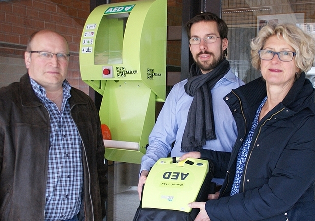 Othmar Zimmermann (links) hat den Kasten per Knopfdruck geöffnet. Die Liegenschaftenvorsteher Beatrice Sommerauer (politische Gemeinde) und Martin Wälti (Sekundarschulgemeinde) zeigen den AED. (Bild Thomas Stöckli)